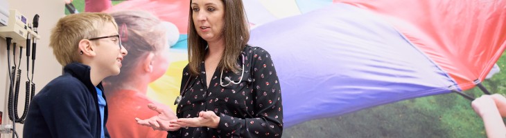 Physician talks to child sitting in clinial room, a picture of children playing outside appears on the wall