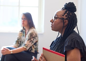 A Black woman with long braided hair holds a folder while seated in a bright room, with another woman seated farther in the background near a window.